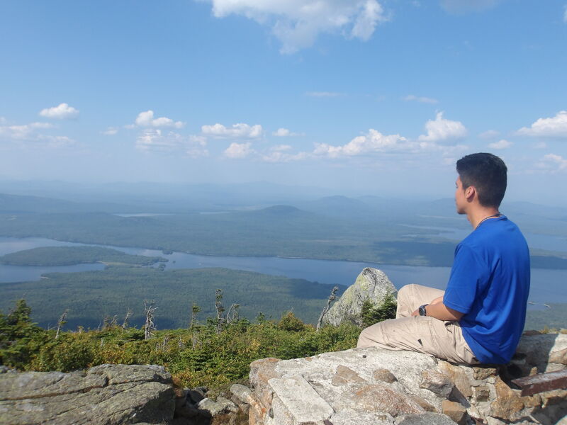 The image shows a man sitting on a rock, looking out at a scenic view. The landscape includes a lake, mountains, and a blue sky with clouds. The man is wearing a blue shirt and khaki pants. He seems to be enjoying the view.